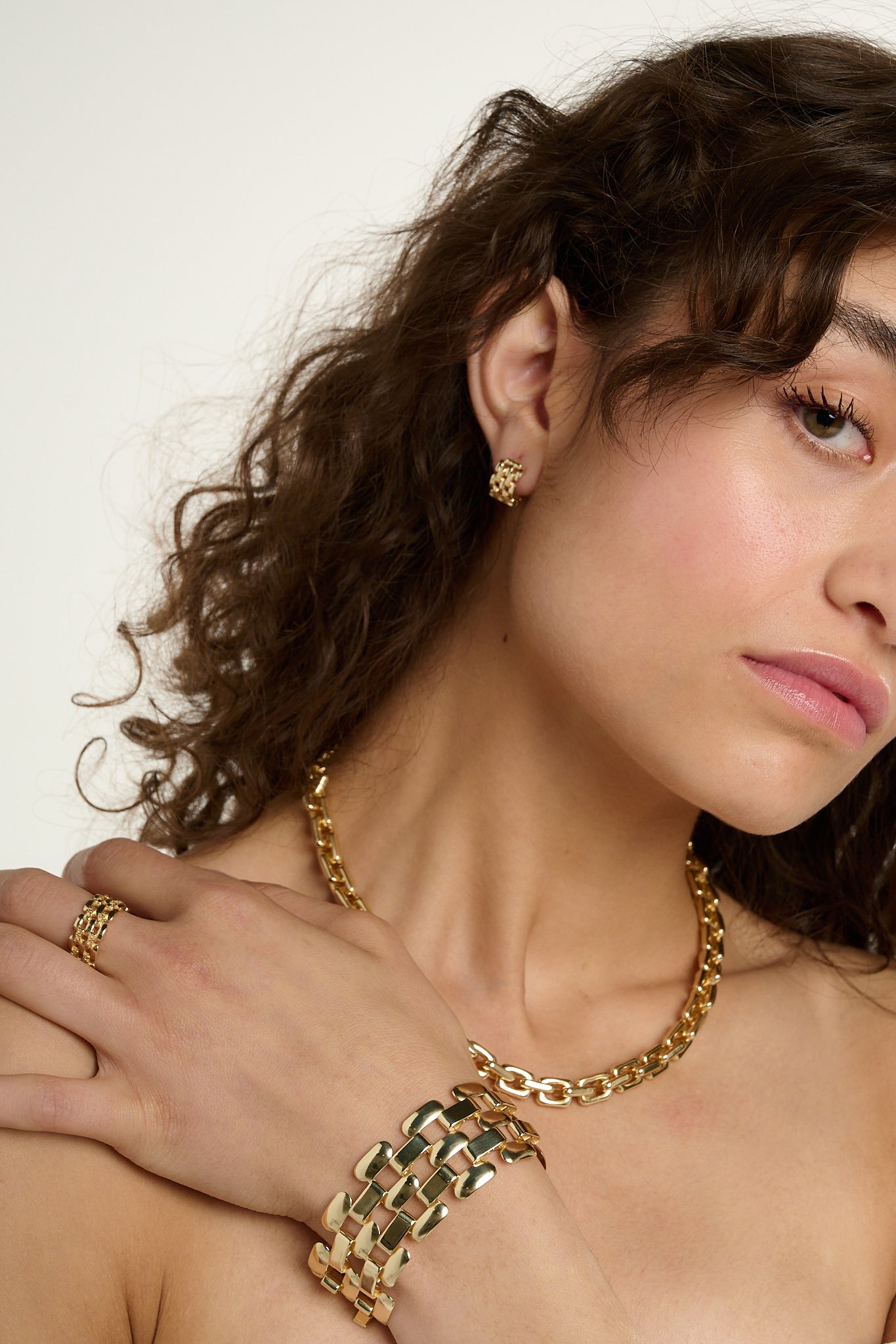 Woman wearing gold jewelry including earrings, necklace, and bracelet against a neutral background